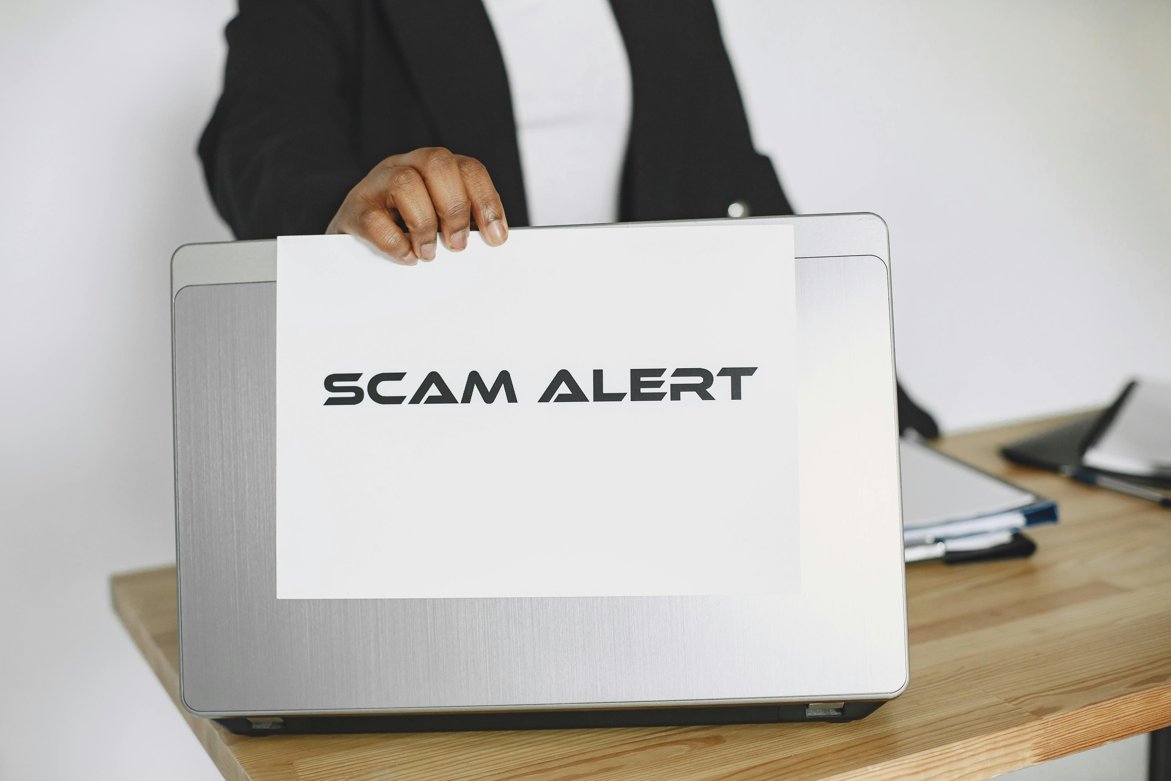 A laptop on a desk with a hand holding a scam alert warning sign